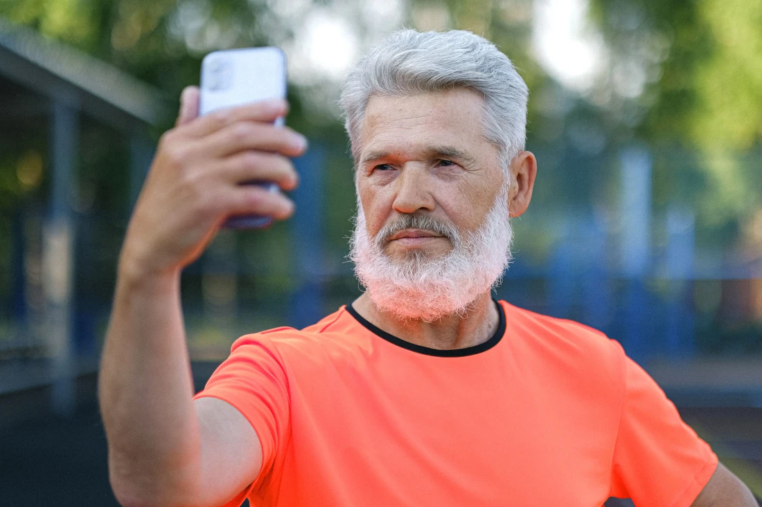 Homem de barba branca e camiseta laranja tirando uma selfie com o celular, com um semblante tranquilo, em um ambiente esportivo ao ar livre.