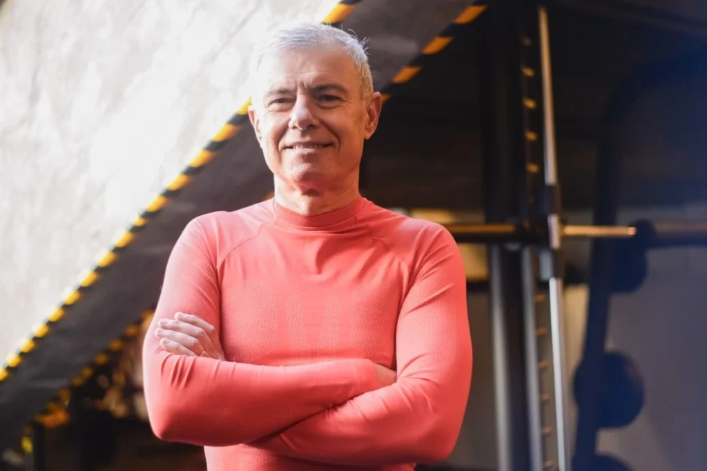 Homem de 60 anos, com cabelos curtos e grisalhos, vestindo uma camisa esportiva vermelha de manga longa, de braços cruzados e sorrindo com confiança em uma academia.