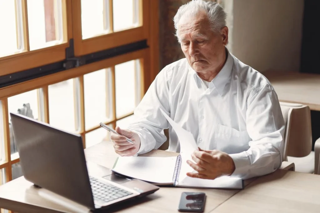 Homem de 60 anos, com cabelos brancos e camisa social, sentado à mesa de madeira com um notebook aberto, lendo atentamente um documento em suas mãos.