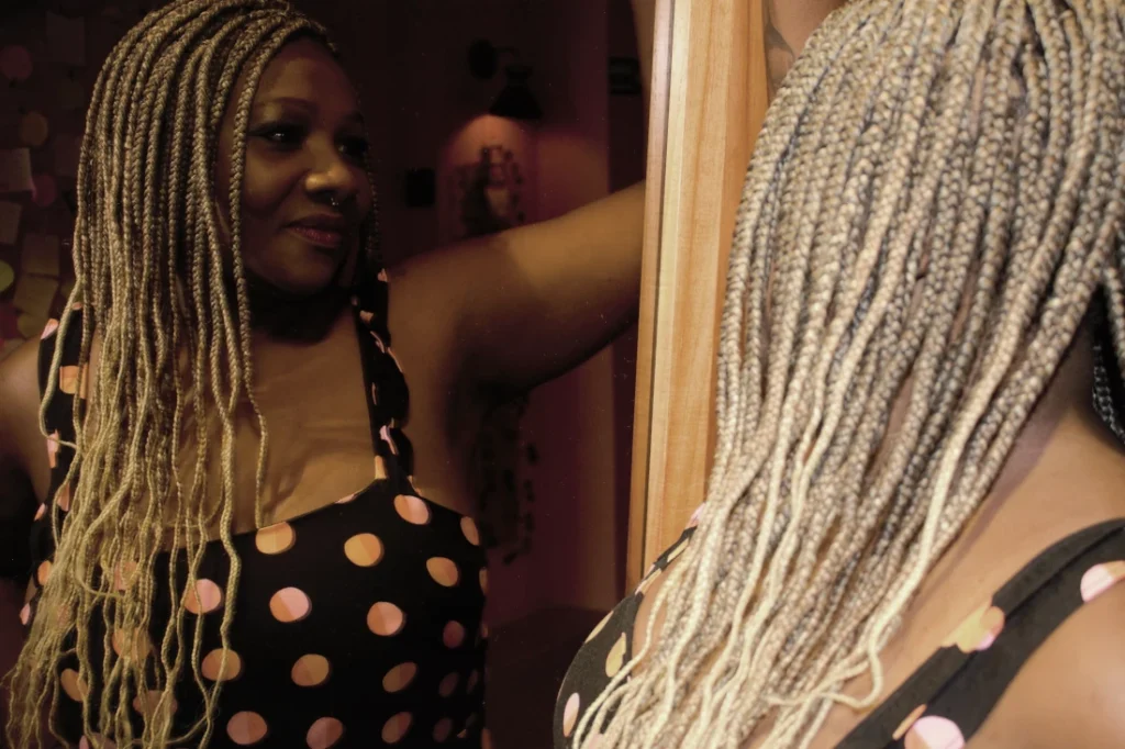 Mulher madura negra, com cabelos trançados longos e blusa preta com bolinhas coloridas, olhando para o próprio reflexo no espelho com um semblante de aceitação.