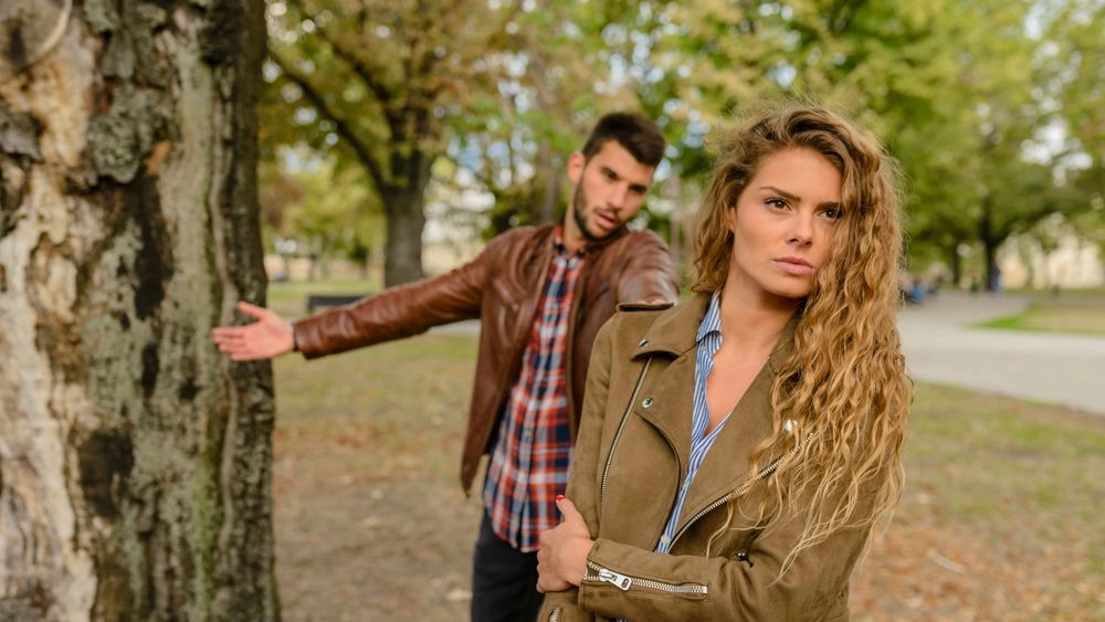 Homem discutindo de forma agressiva com uma mulher de braços cruzados e expressão de exaustão em um parque.