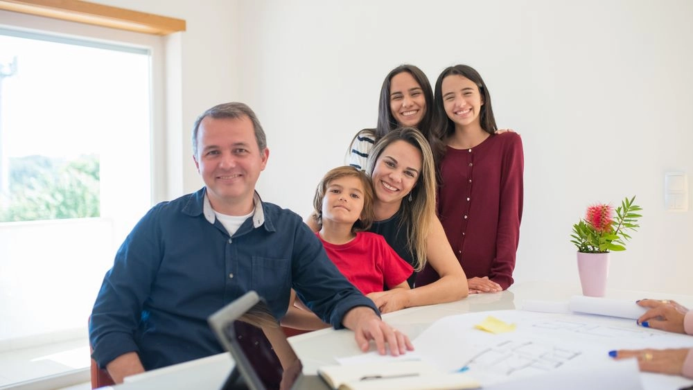 Um pai sorridente sentado em uma mesa clara com um menino pequeno, ao lado de sua esposa e duas filhas adolescentes que também sorriem para a foto.