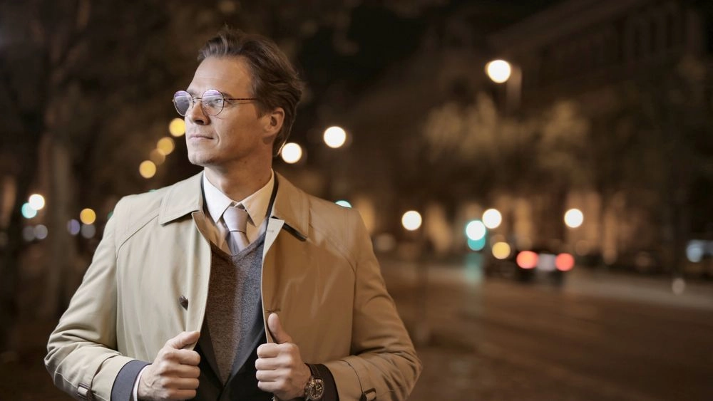 Homem branco, elegante, usando óculos e sobretudo, posicionado em uma rua de cidade iluminada à noite com uma postura imponente e respeitável.