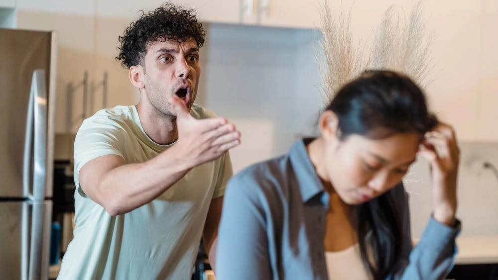 Homem desfocado ao fundo gritando agressivamente e apontando o dedo, enquanto uma mulher em primeiro plano olha para baixo, exausta e segurando a cabeça.