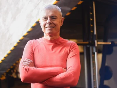 Homem de 60 anos, com cabelos curtos e grisalhos, vestindo uma camisa esportiva vermelha de manga longa, de braços cruzados e sorrindo com confiança em uma academia.