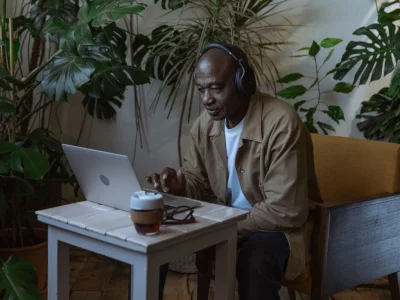 Homem negro maduro usando fones de ouvido e trabalhando em um notebook, sentado em uma cadeira amarela confortável cercado por várias plantas domésticas.